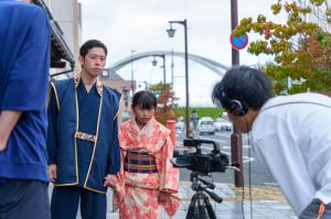 明智光善と明智ひかりの広小路での撮影シーン。二人が道のそばで何かを見つめている様子。手前には撮影スタッフ。