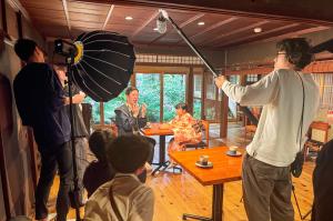 飲食店の店内での撮影の様子。慌てた様子の光善と向かいに座っているひかりを、数名の撮影スタッフが取り囲んでいる。