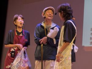 ステージの上でピンマイクをつけて一人の高齢男性と二人の女性が話している。男性は防止をかぶるなどお出かけ中のようであり、2人の女性はエプロン姿だ。