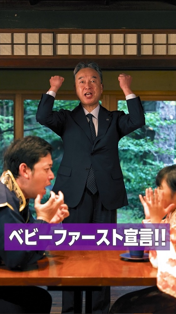 ベビーファースト宣言編1話の市長初登場シーン。飲食店で向かい合って座る女の子とお父さんの間に市長が両手を挙げて立ち、「ベビーファースト宣言」と叫んでいる。